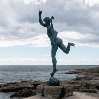 Staty av en dansande pojke i brons som balanserar på en fot vid havet, med ena handen uppsträckt mot himlen och klippor i bakgrunden.