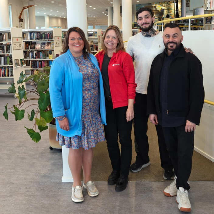 Fyra personer står tillsammans och ler i ett bibliotek med bokhyllor i bakgrunden.