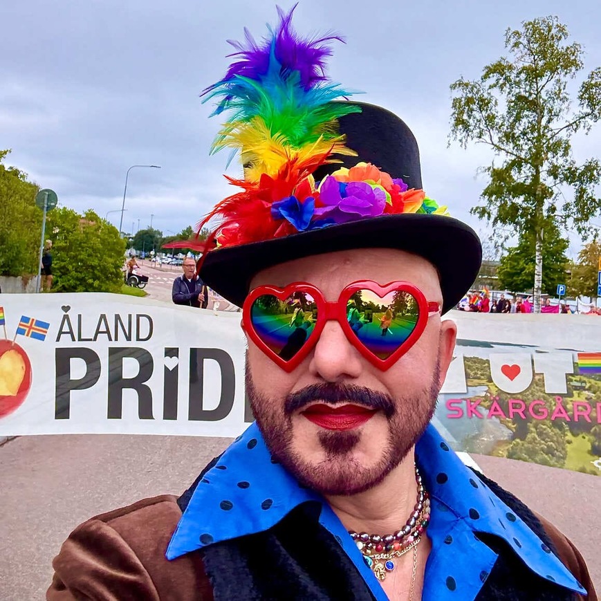 En person med färgglada accessoarer och regnbågsfärgade glasögon står framför en skylt med texten ‘Åland Pride’