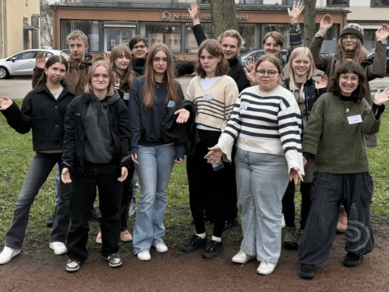 En grupp ungdomar står utomhus och poserar tillsammans framför en byggnad, flera av dem vinkar mot kameran.
