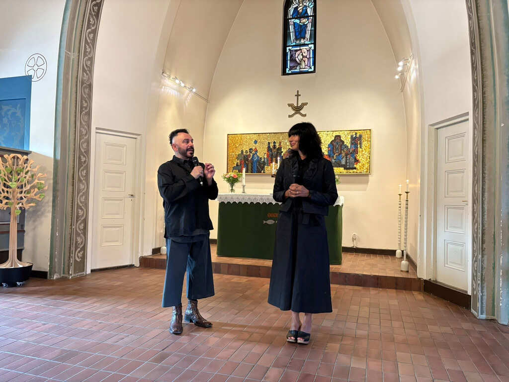 Två personer står framför altaret i en kyrka och talar till publiken under en utställning.