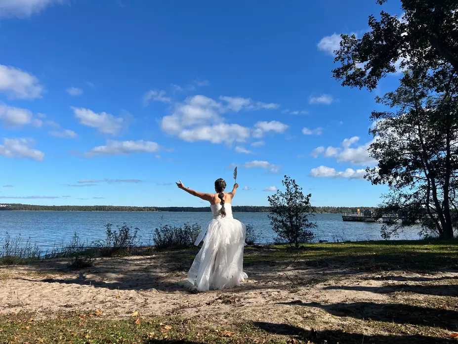 Person i ljus klänning står på en sandstrand med ryggen mot kameran och armarna utsträckta mot havet, under en klar blå himmel med spridda moln och träd som ramar in kustlandskapet.