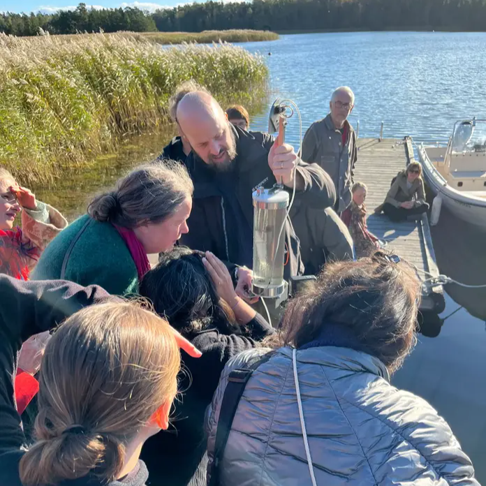 n grupp människor samlas på en brygga vid vattnet medan en person visar upp ett vattenprov i ett genomskinligt kärl.