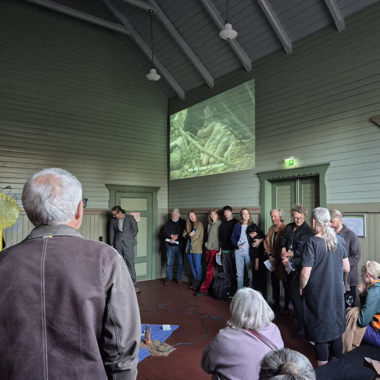 Grupp människor står samlade i ett rum kring en installation med projektion på väggen, där ett videoverk visas ovanför dem.