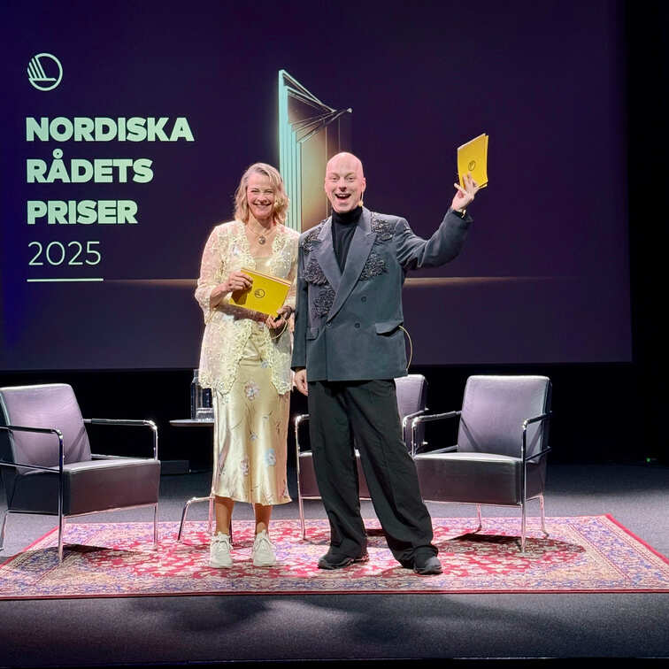 Två värdar står på scenen och håller upp programkort medan de ler mot publiken, med Nordiska rådets priser 2025 projicerat bakom dem.