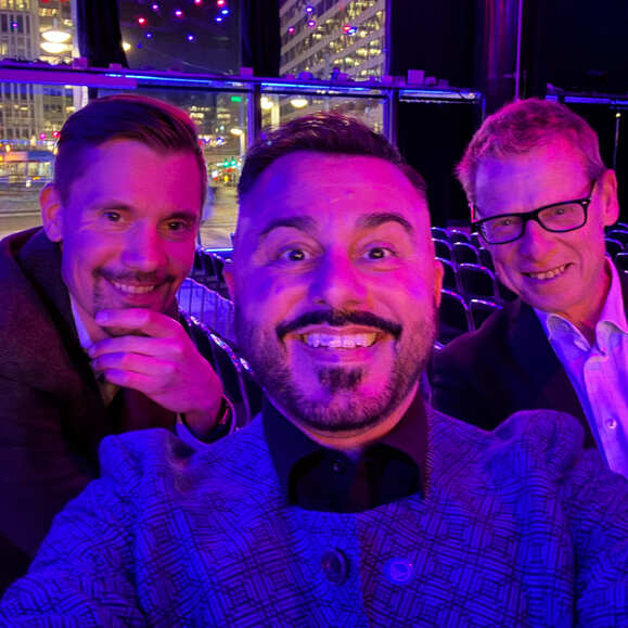 Tre personer tar en selfie i en upplyst sal med scen och publikplatser i bakgrunden.