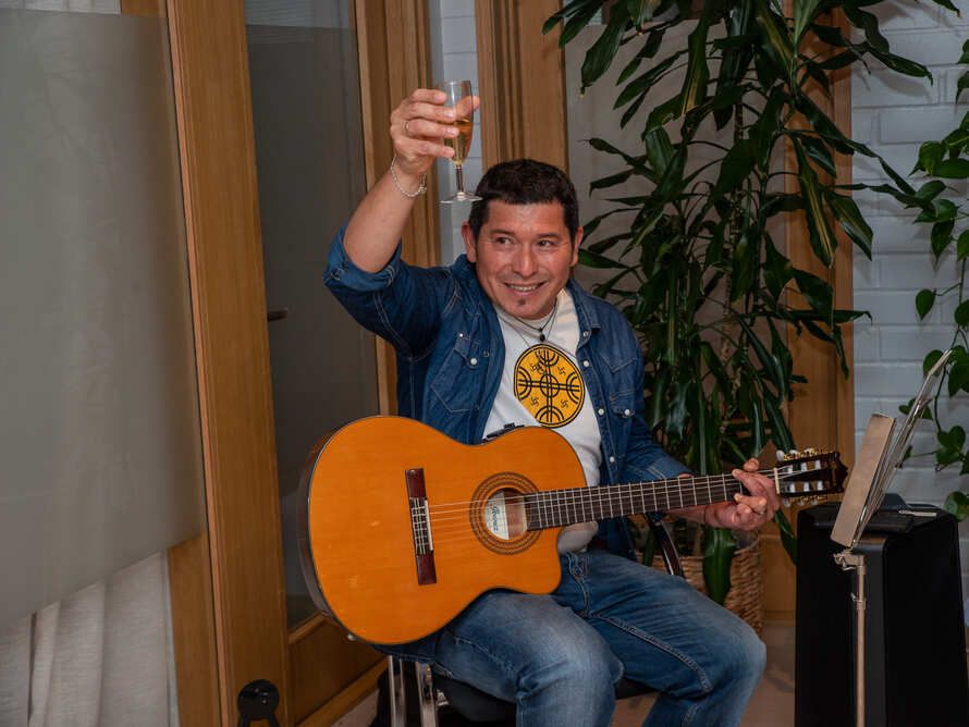 En person sitter med en gitarr och höjer ett glas i en informell musikstund.